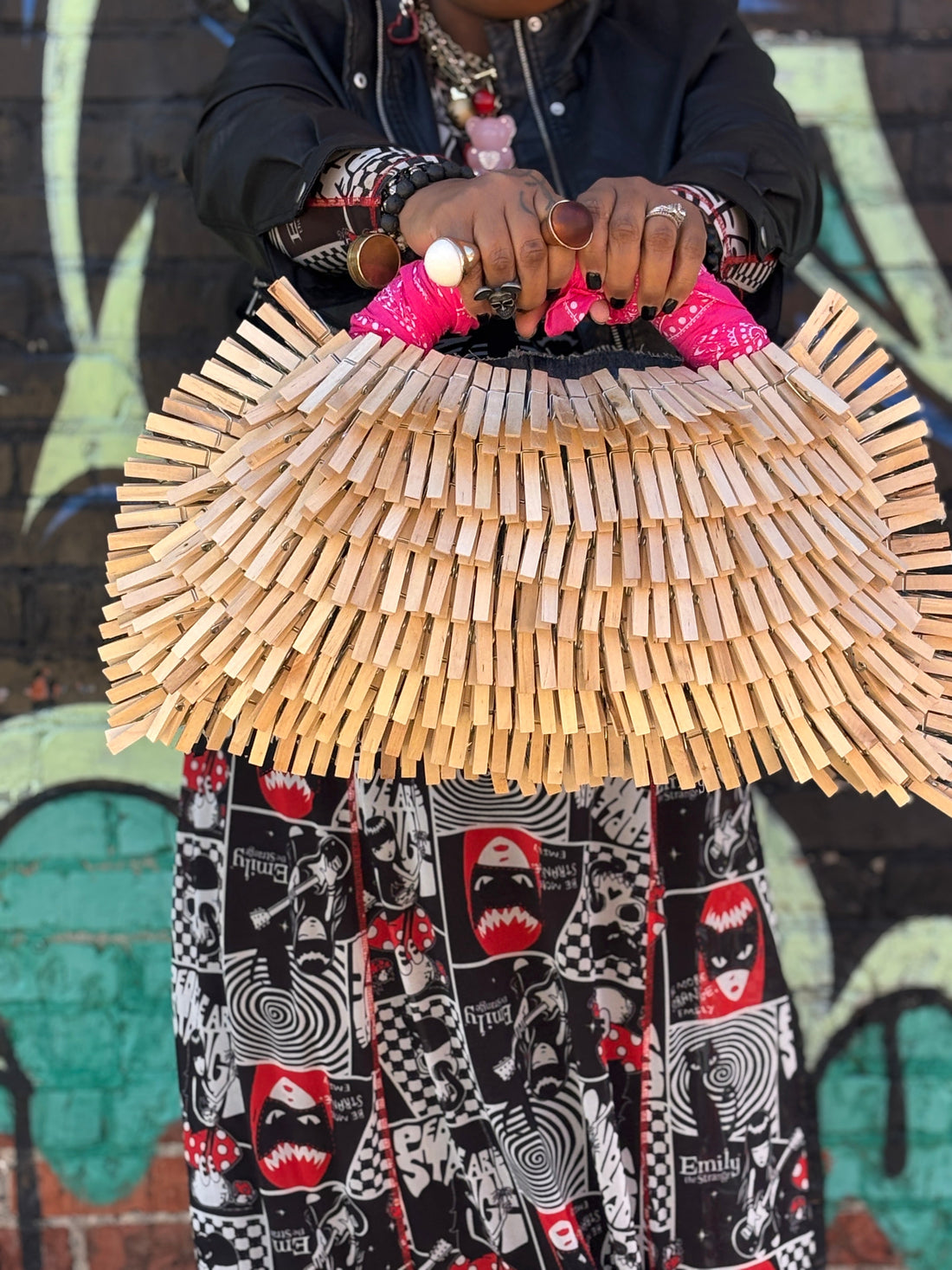 Woman holding a funky handmade clothespin bag with a pink bandanna handle, standing in front of a graffiti wall, wearing bold accessories and expressive personal style
