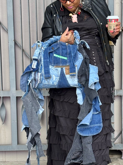 Person holding a denim jacket with patches, wearing a layered outfit, and holding a coffee cup.